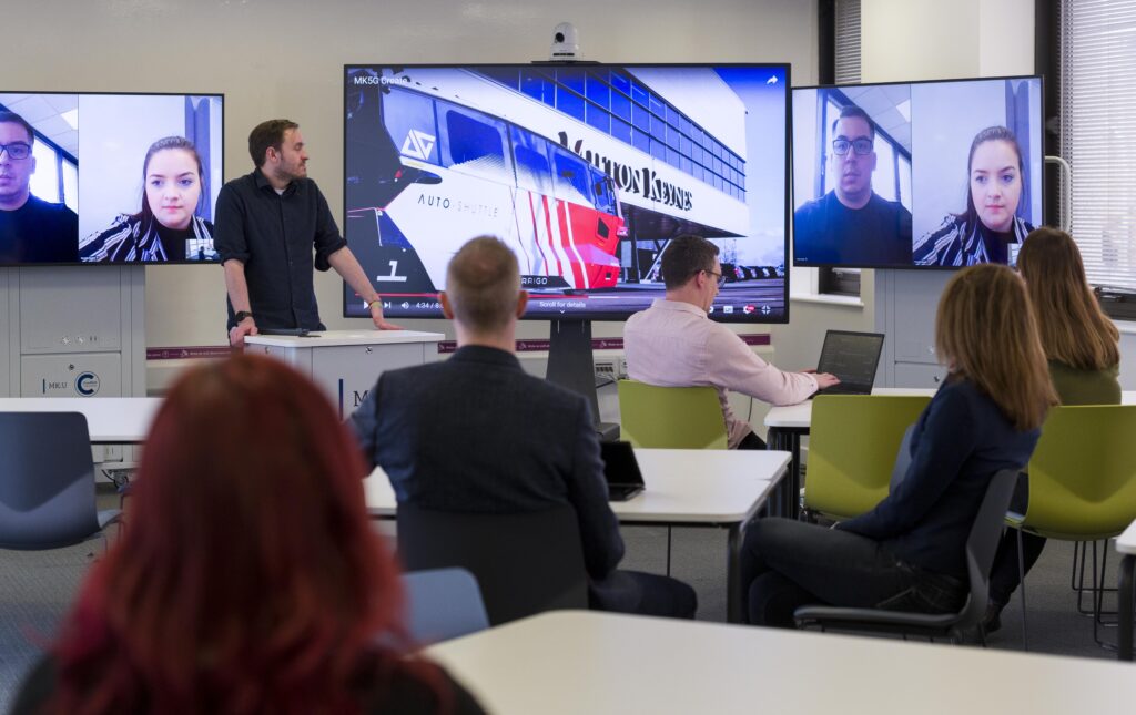 Classroom presentation with remote participants shown on video screens.