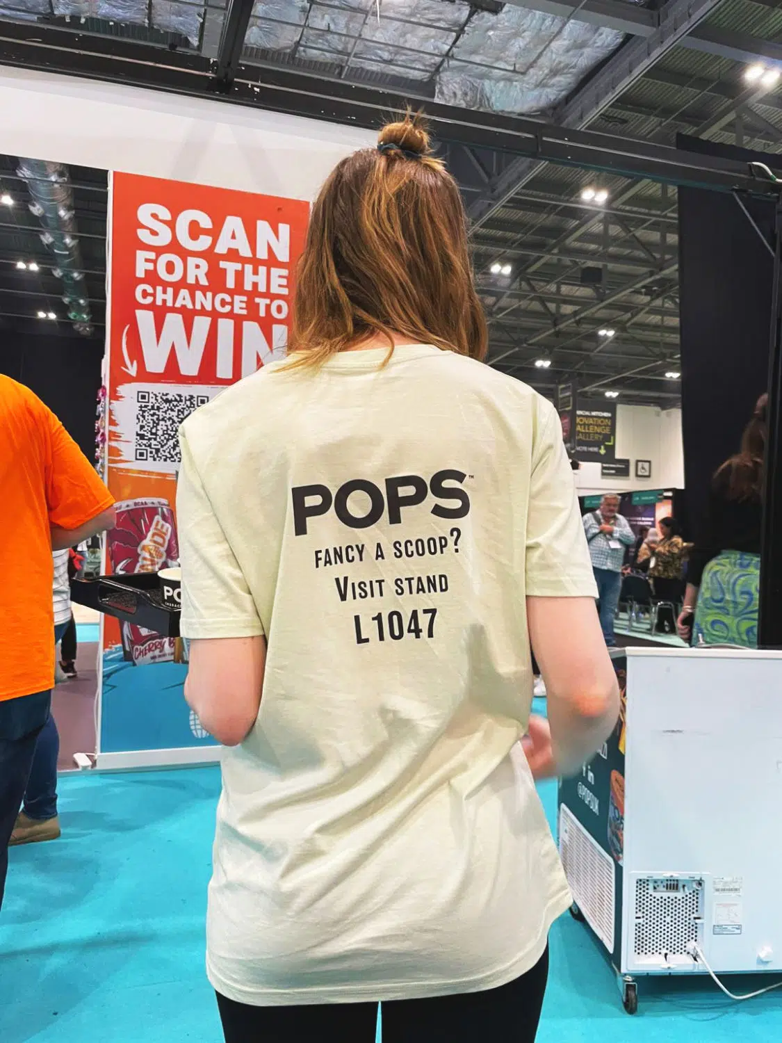 The back of a person wearing a pale yellow t-shirt with the "POPS" logo and text inviting attendees to "VISIT STAND L1047" at an event.
