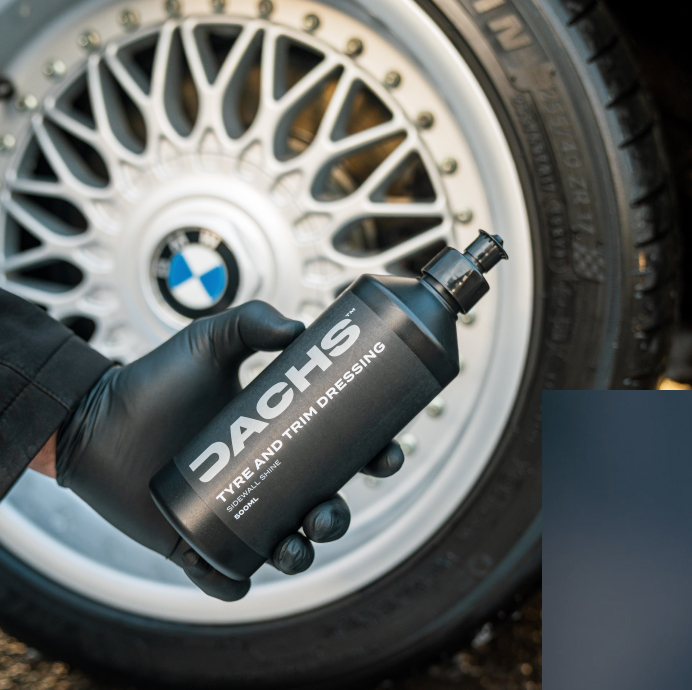 A hand in a black glove holding a bottle of Dachs Tyre and Trim Dressing in front of a clean alloy wheel.