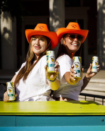 two woman wearing orange cowboy hats holding cans of malibu cocktail